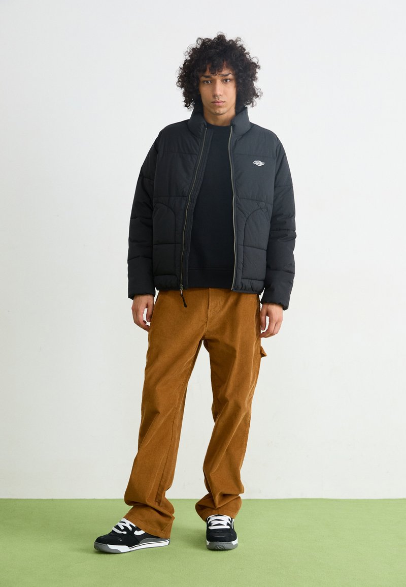 Doudoune noire avec un design matelassé et un col montant, associée à un pantalon en velours côtelé de couleur rouille et des baskets noires avec des accents blancs.