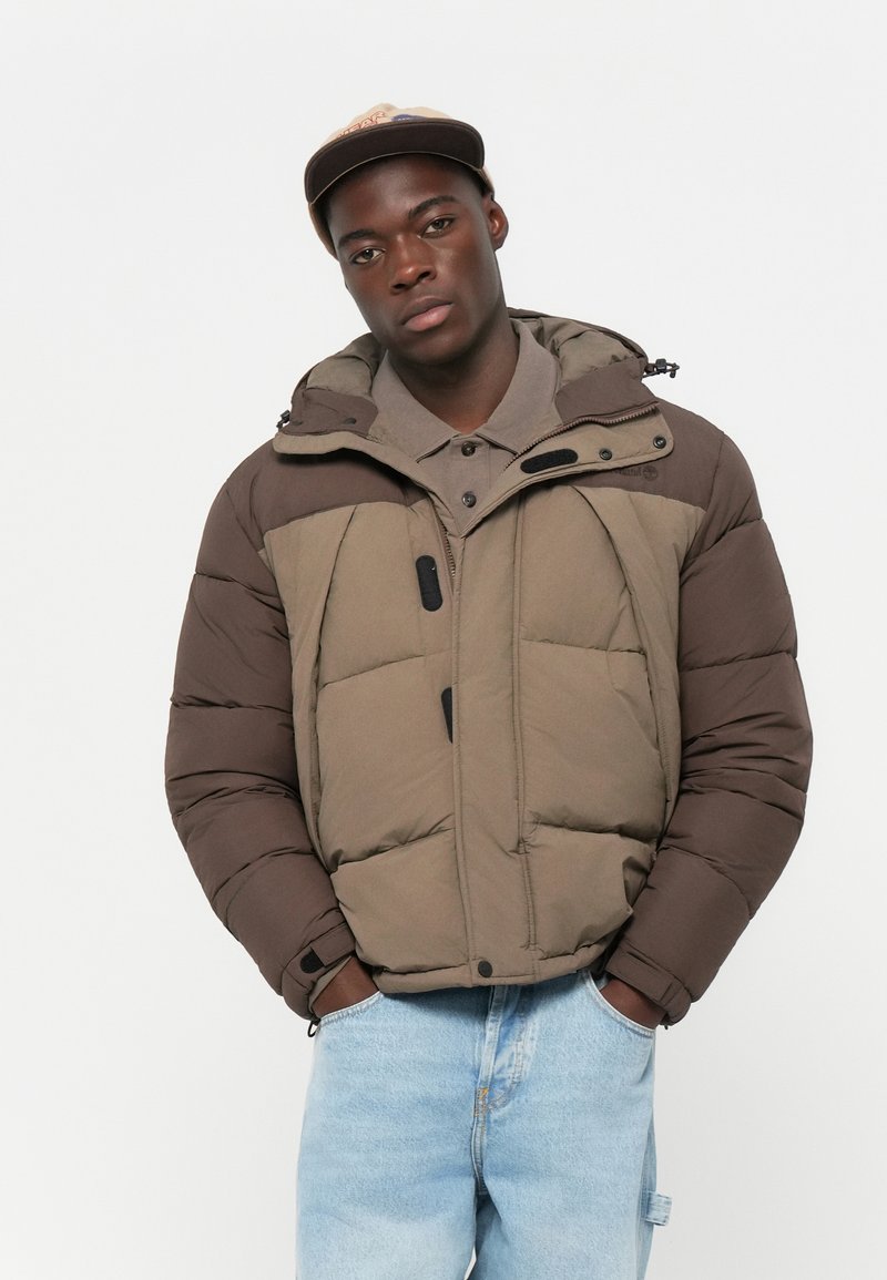 Chaqueta tipo puffer en tonos marrones y verde oliva con capucha, que cuenta con cierre de cremallera, botones a presión y una textura de tela con grandes secciones acolchadas.