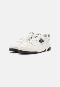 White athletic sneakers with black accents, textured leather upper, perforations, flat black rubber sole, and lace-up closure.
