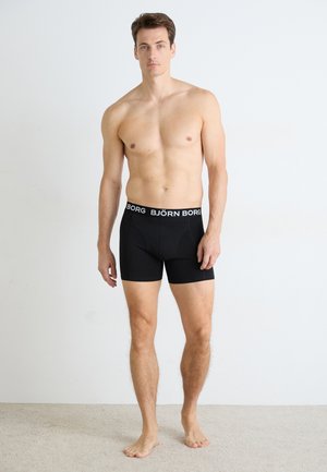 Männliches Modell, das barfuß gegen eine weiße Wand steht und schwarze Björn Borg Boxershorts mit einem markierten Bund trägt.