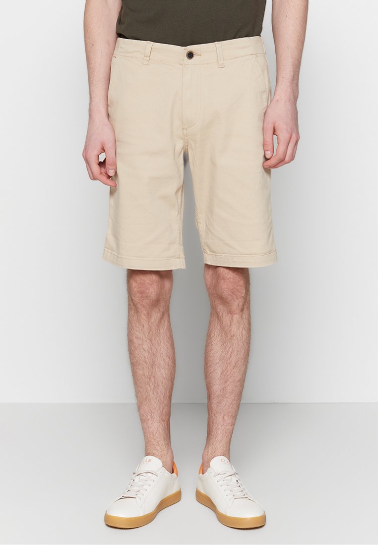 Homme portant un short beige jusqu'au genou, des baskets blanches avec semelles en gomme et une chemise vert foncé, debout devant un fond uni.