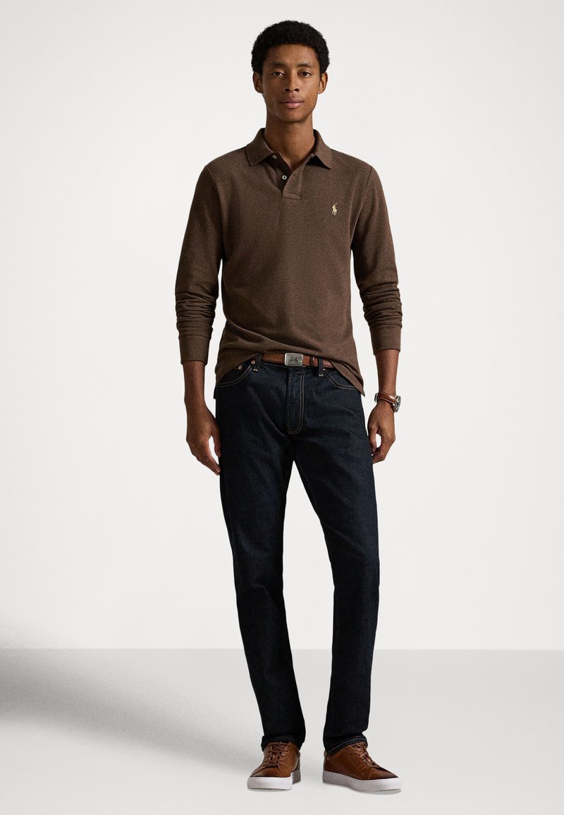 Braunes langärmeliges Poloshirt mit Kragen, dunkle Jeans und braune Turnschuhe. Das Shirt verfügt über ein dezentes Logo und einen strukturierten Stoff.