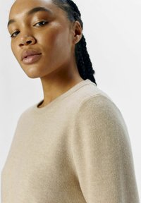 Pull beige avec un col rond, texture douce et coutures minimales. Le tissu semble léger et lisse, idéal pour superposer.