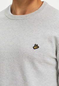 Grå texturerad tröja med rund halsringning. Har en liten gul och svart logotyp på bröstet. Materialet verkar mjukt och stickat.