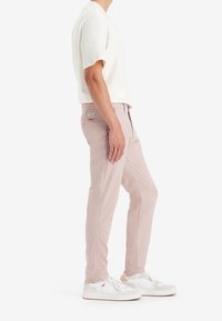 Ljusrosa byxor med smal passform, sidofickor, etikett bak, i kombination med vita sneakers och en krämfärgad kortärmad skjorta.