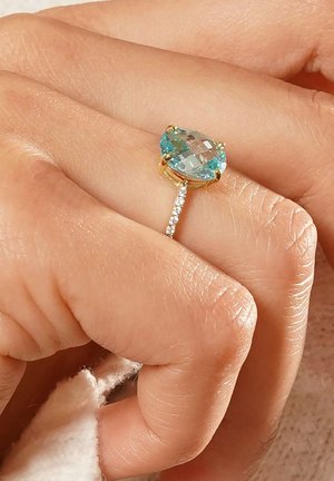 Goldring mit einem ovalen, hellblauen Edelstein mit Facetten, begleitet von einem Band, das mit kleinen klaren Steinen verziert ist. Hautfarbe sichtbar.