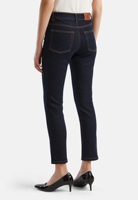 Tmavě modré úzké džíny s oranžovým stehem, dvěma zadními kapsami a hnědou koženou etiketou na pase, doplněné černými vysokými podpatky.