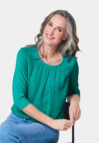 Groene, geplooide blouse met driekwart mouwen, ronde hals, gemaakt van lichtgewicht stof. Gecombineerd met blauwe jeans, zittend, neutrale achtergrond.