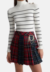 Hvid ribbet turtleneck sweater med sorte striber, parret med en rød og marineblå skotsk ternet plisseret nederdel med en kædeaccessory og dekorative knapper.