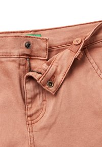 Pantalones de mezclilla de color rosa claro con cierre de botón, cremallera y detalles de costura. Presentan un bolsillo frontal y una etiqueta de marca verde.