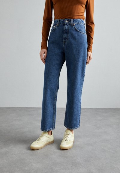 Hochgeschnittene blaue Denim-Jeans mit geradem Bein, ausgestattet mit einem Knopf- und Reißverschluss und kontrastierenden Nähten. Kombiniert mit hellen Sneakers.