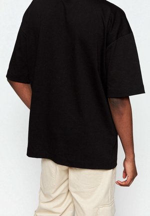 Osoba ubrana w luźną czarną koszulę z krótkim rękawem i jasnobeżowe spodnie, pokazana od tyłu, z prawą ręką swobodnie opuszczoną wzdłuż ciała.