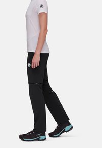 Persoon met wit shirt met korte mouwen, zwarte afritsbare wandelbroek en zwart-blauwe wandelschoenen, staand in profiel tegen een witte achtergrond.