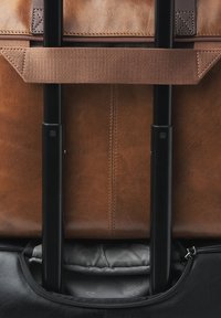 Braune Ledertasche mit schwarzem, ausziehbarem Griff, ausgestattet mit einem Reißverschluss und weichem grauem Innenfutter. Genähte Akzente auf dem Leder.