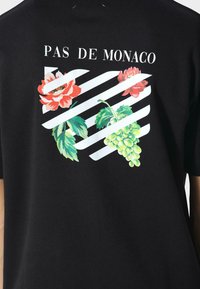 Czarna koszula w białe ukośne paski, czerwone kwiaty, zielone winogrona oraz napis "PAS DE MONACO" na plecach.