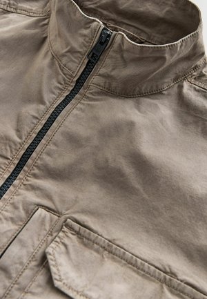 Pruun lukuga jope kõrge kraega, millel on must lukk, esi tasku ja sile tekstuur. Materjal näib kerge ja vastupidav.