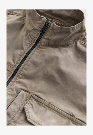 Pruun lukuga jope kõrge kraega, millel on must lukk, esi tasku ja sile tekstuur. Materjal näib kerge ja vastupidav.