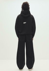 Personne avec des dreadlocks vue de dos, portant un sweat à capuche noir avec un logo blanc et un jean noir à jambes larges, sur un fond uni.