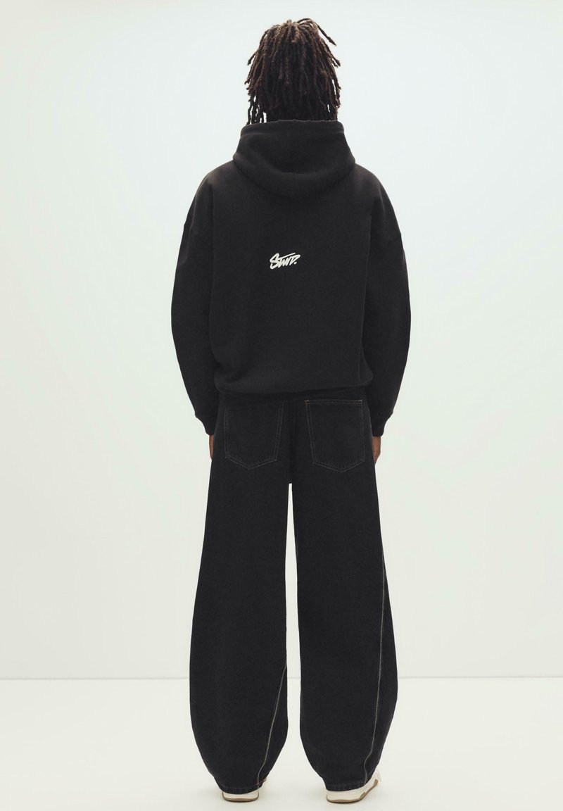 Personne avec des dreadlocks vue de dos, portant un sweat à capuche noir avec un logo blanc et un jean noir à jambes larges, sur un fond uni.