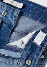 Interior de unos vaqueros de denim con un color azul, costuras visibles, cremallera, botón a presión y etiqueta de cuidado del tejido. Superficies lisas y texturizadas.