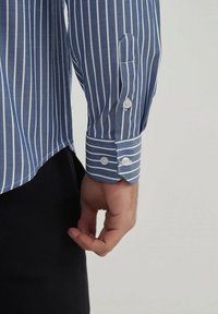 Bras droit vêtu d'une chemise à manches longues verticalement rayée bleue et blanche avec un poignet boutonné, reposant à côté d'un pantalon noir devant un fond uni.