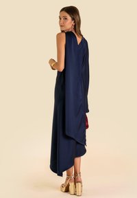 Navyblaues Satinkleid mit drapiertem Rücken, ärmellosem Design, asymmetrischem Saum und goldenen Plateau-Absätzen. Mit einer roten Clutch accessoriert.