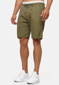 Shorts vert olive en tissu texturé, avec une taille à cordon de serrage réglable et des poches latérales, associés à des chaussures de sport blanches.