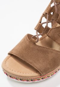 Gros plan d'une sandale en daim marron à bout ouvert avec des lacets croisés et un détail tissé multicolore sur le bord de la semelle intermédiaire.