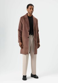 Bruine, zachte stoffen jas met een reverskraag, twee knopen en zijzakken, gecombineerd met lichtbeige broek en zwarte schoenen.