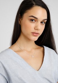 Sølv halskæde med en lille bogstav "M" vedhæng, vist på en person iført en lyseblå V-hals top. Glat tekstur, minimalistisk design.