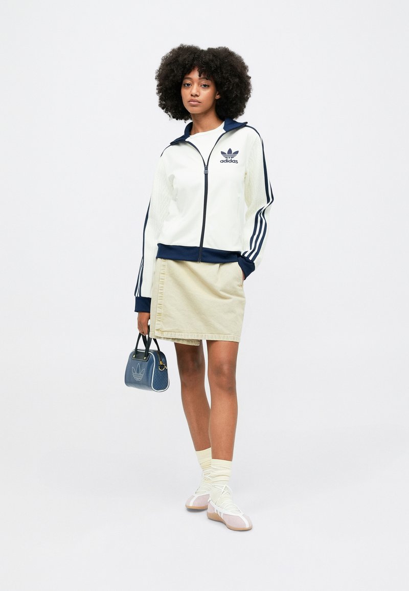 White zip-up jacket with navy accents and three stripes, beige skirt, blue handbag, cream socks, and light pink shoes.