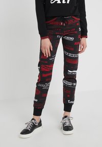 Czarne legginsy z nadrukiem przedstawiającym różne czerwone logo i napisy związane z podróżami. Wykończone czarnymi mankietami przy kostkach oraz sznurkiem w pasie.