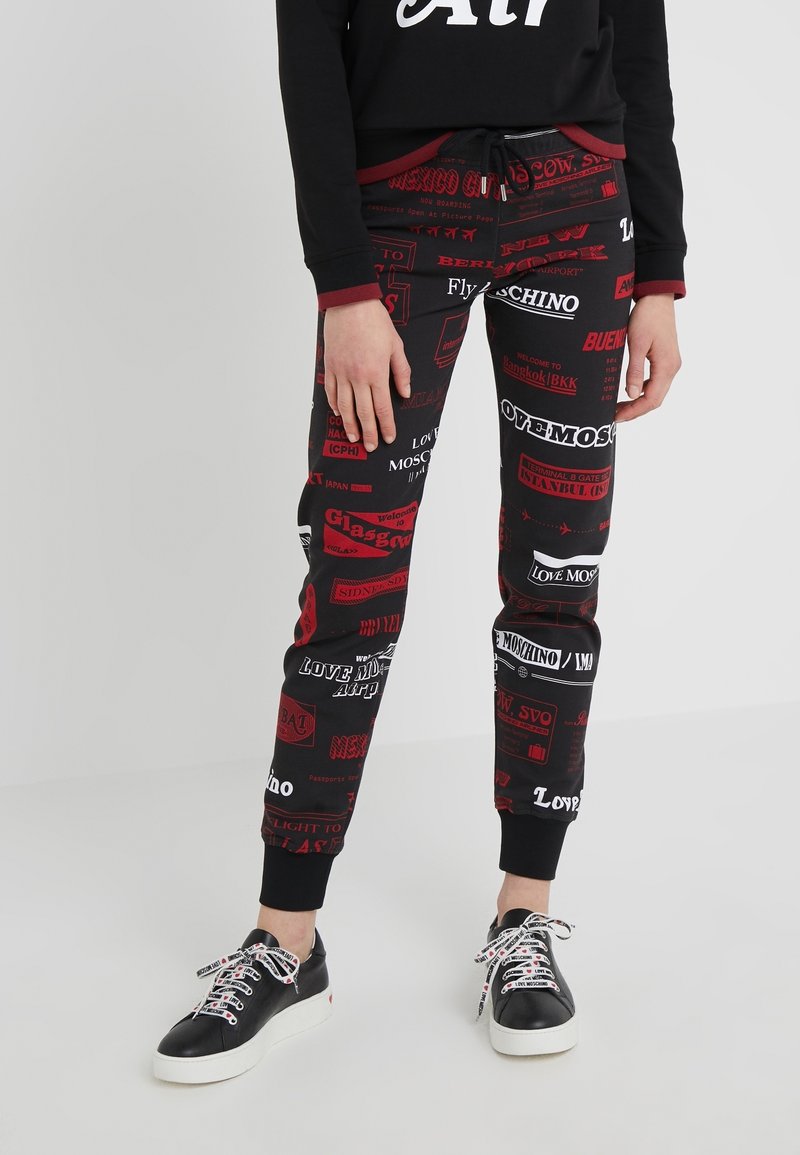 Czarne legginsy z nadrukiem przedstawiającym różne czerwone logo i napisy związane z podróżami. Wykończone czarnymi mankietami przy kostkach oraz sznurkiem w pasie.