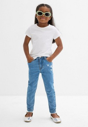 Ragazzina con occhiali da sole verdi, t-shirt bianca, jeans blu con piccoli topolini bianchi e ballerine argentate, mani in tasca.