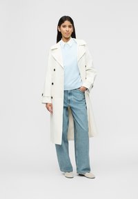 Femme debout portant un trench-coat blanc, un pull bleu clair par-dessus une chemise blanche à col, un jean bleu ample et des chaussures de couleur claire.