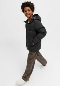 Giubbotto nero imbottito con cappuccio, cerniera frontale e tasche laterali, indossato sopra una camicia chiara, abbinato a pantaloni di velluto a coste marroni e sneakers bianche.