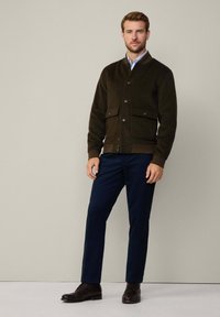 Dark green bomber jacket with button closure, two front pockets, ribbed cuffs, paired with navy trousers and brown leather shoes.