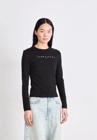 Crna majica dugih rukava s okruglim izrezom i bijelim logotipom "TOMMY JEANS" na prsima; uparena s svijetloplavim trapericama.