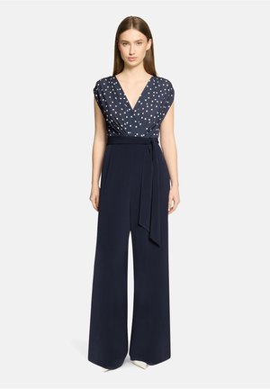 Frau steht und trägt einen marineblauen Overall mit einem ärmellosen Oberteil mit weißen Punkten, tiefem V-Ausschnitt, weitem Bein und passendem Stoffgürtel, der in der Taille gebunden ist.