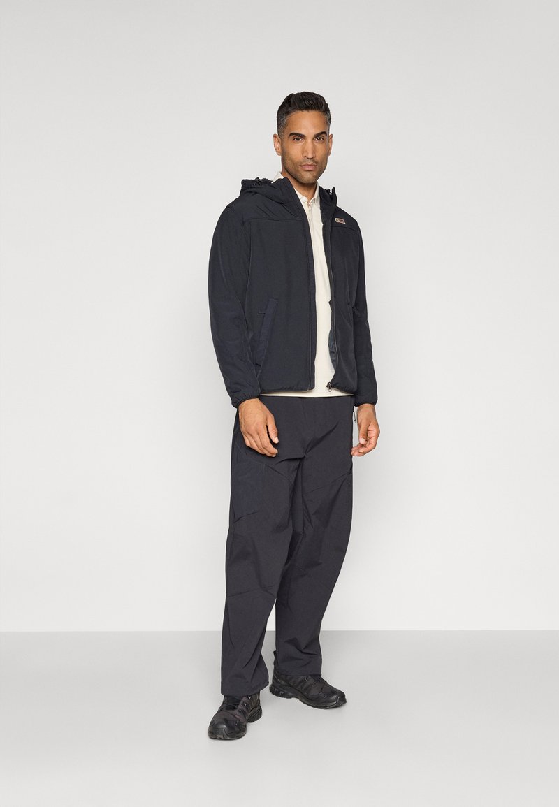 Veste à capuche noire pour homme avec une texture lisse, dotée de poches avant et d'une fermeture éclair, associée à un pantalon noir ample et des chaussures noires.