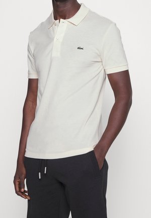 Poloshirt aus hellbeigem Baumwollstoff mit einer Knopfleiste mit zwei Knöpfen, kurzen Ärmeln und einem grünen gestickten Logo auf der Brust.