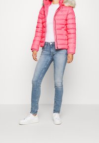 Růžová prošívaná bunda s kapucí lemovanou kožešinou, s obrysovým prošíváním, nošená přes bílý top a doplněn�á světle modrými úzkými džínami a bílými teniskami.