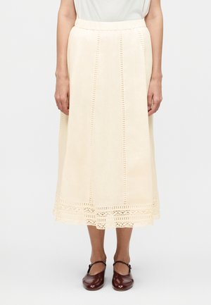 Žena nosi krémovú midi sukňu s vertikálnymi detailmi s očkami a čipkovaným lemom, v kombinácii s hnedými topánkami Mary Jane.