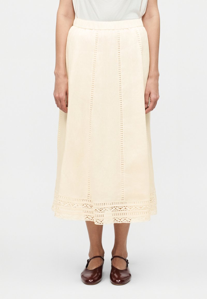 Γυναίκα που φοράει φούστα midi εκρού με κάθετες λεπτομέρειες από τρύπες και δαντελένιο τελείωμα, σε συνδυασμό με καφέ παπούτσια Mary Jane.