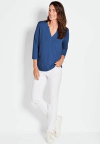 Blaue V-Ausschnitt-Bluse mit drei Viertelärmel und abgerundetem Saum, kombiniert mit weißen Straight-Leg-Hosen und weißen Turnschuhen. Einfaches Design, lockere Passform.