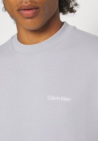 Camiseta gris claro con cuello redondo acanalado y el logo blanco de "Calvin Klein" en el pecho, fabricada en un suave y ligero material de algodón.