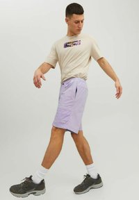 Light purple shorts with an elastic waistband, side pockets, and logo detail. Beige t-shirt features a graphic print. Gray sneakers with white socks.