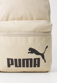 Bežový látkový batoh s čiernym logom PUMA a zipsom na prednej strane. Textúrovaný povrch s pevným tvarom.