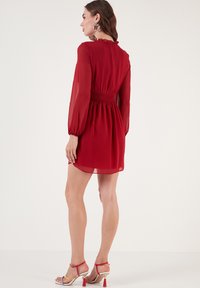 Rode chiffon jurk met lange, opgeblazen mouwen, smocktail en gerimpelde halslijn. Draagt met gekruiste, veelkleurige hoge hakken sandalen.