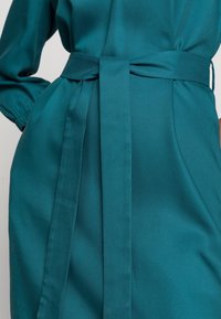 Blågrøn kjole med taljebælte, glat tekstur og plisserede detaljer i siden. Lange ærmer med samlede manchetter.